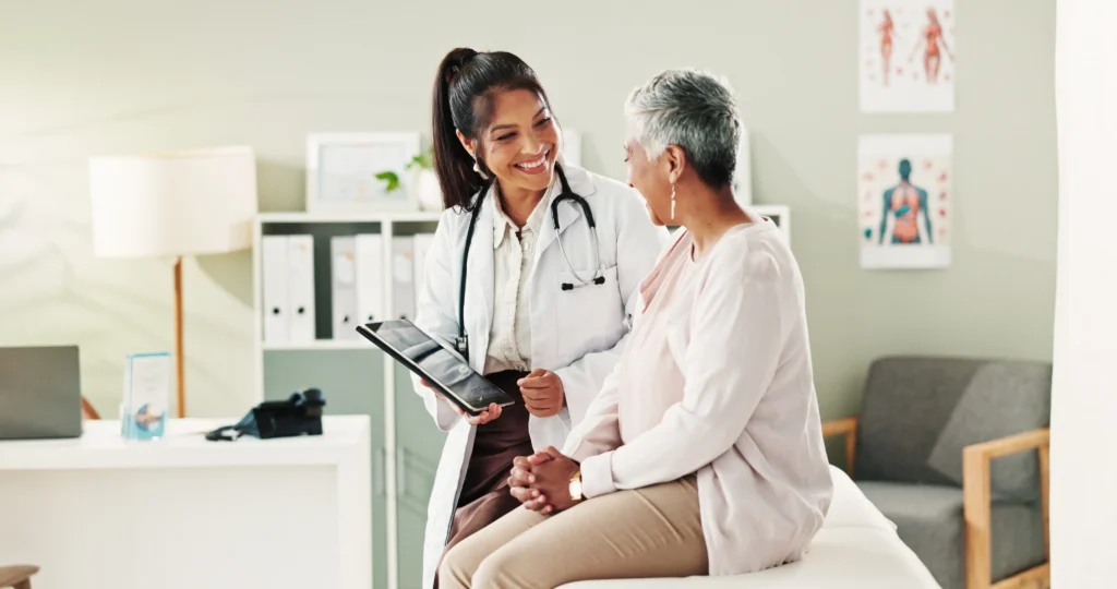 Dentist reviews information on a tablet while speaking with an older patient in an exam room.