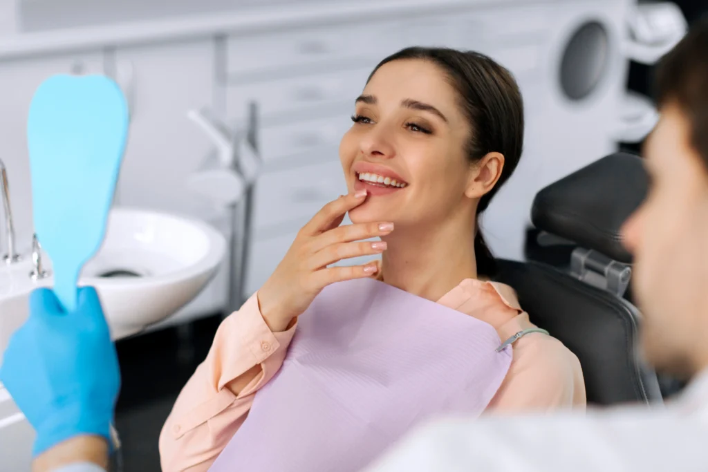 Person in a dental chair smiles while looking into a hand mirror and touching the chin.
